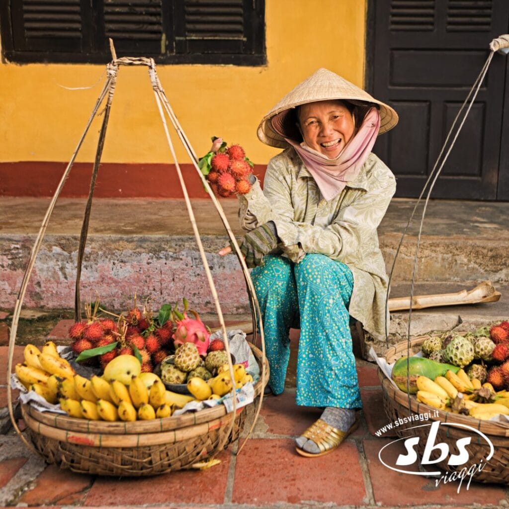 Una donna che indossa un cappello conico è seduta a terra, sorridente, con accanto dei cesti di frutta tropicale: una classica scena di un tour in Vietnam che cattura il fascino vibrante della vita quotidiana.