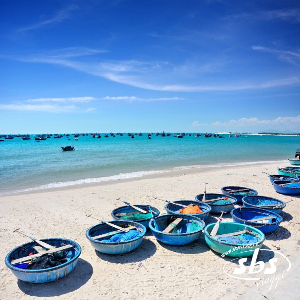 Barche blu con ceste rotonde sono allineate su una spiaggia sabbiosa come parte di un memorabile Tour Experience Vietnam, con barche da pesca che galleggiano nel mare turchese sotto un cielo azzurro e limpido.