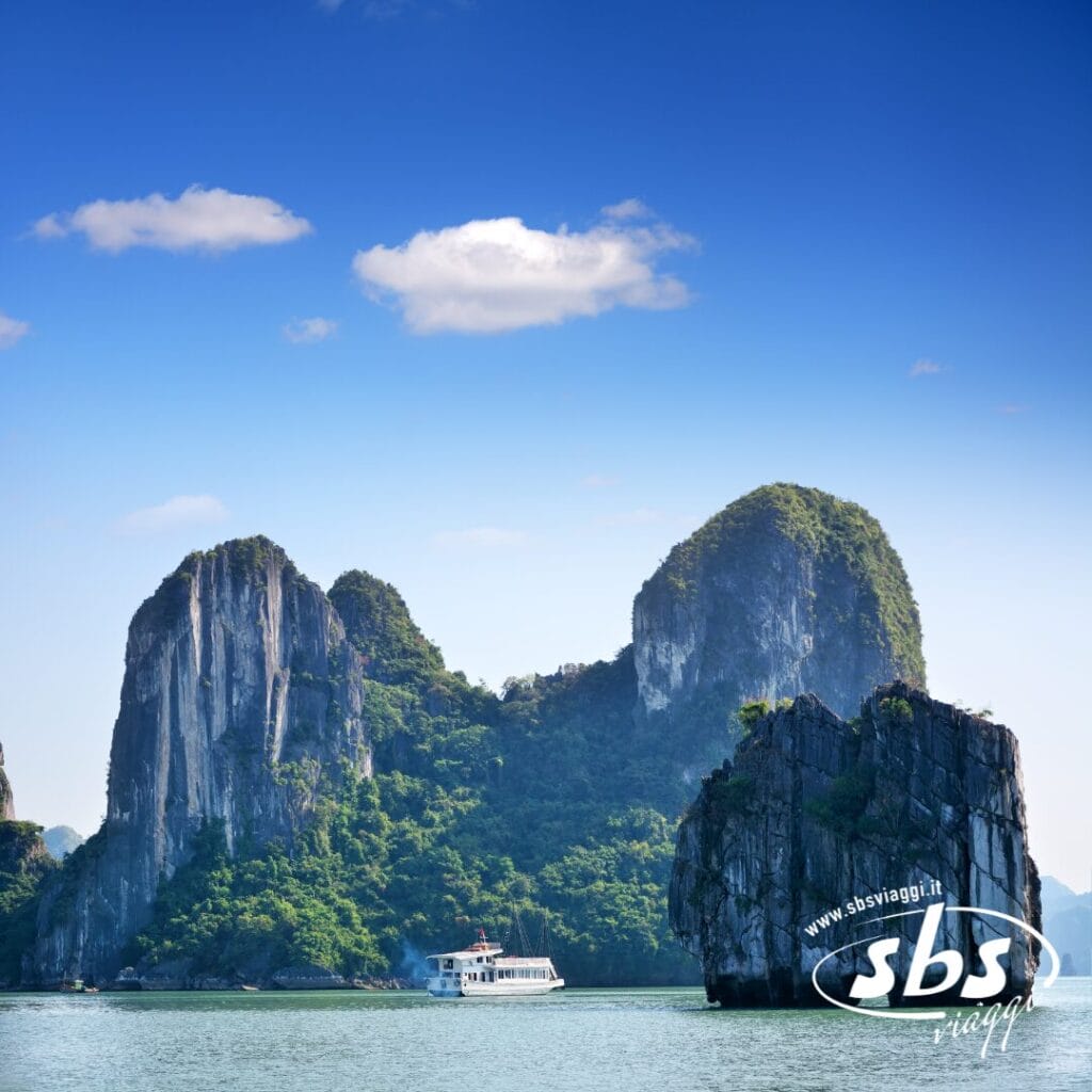 Una barca bianca naviga tra isole calcaree verdi e formazioni rocciose su acque calme e azzurre sotto un cielo limpido: uno scenario indimenticabile di un tour in Vietnam. Il logo