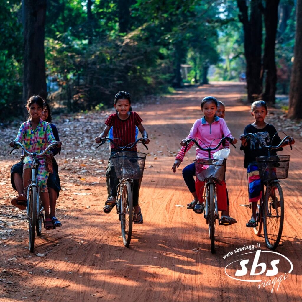 Quattro bambini vanno in bicicletta uno accanto all'altro su una strada sterrata attraverso una zona boscosa, mentre la luce del sole filtra tra gli alberi, catturando lo spirito d'avventura che si prova durante un viaggio Grantour o Tour Cambogia.