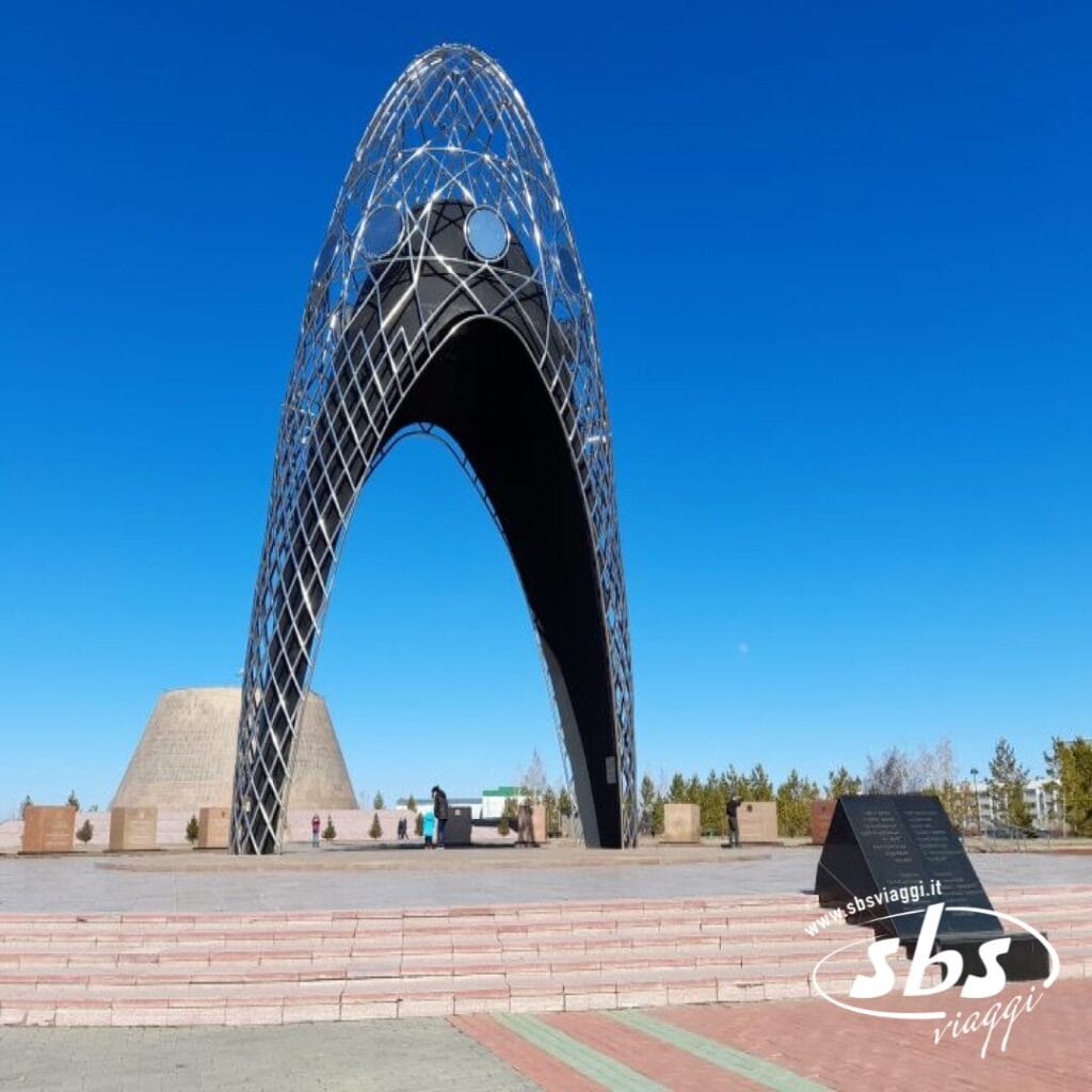 Un grande monumento metallico ad arco si erge su una piazza pavimentata sotto un cielo azzurro e limpido, con alcune persone nelle vicinanze e una targa informativa in primo piano: una tappa obbligata per qualsiasi Tour Kazakistan o viaggio Kazakistan.