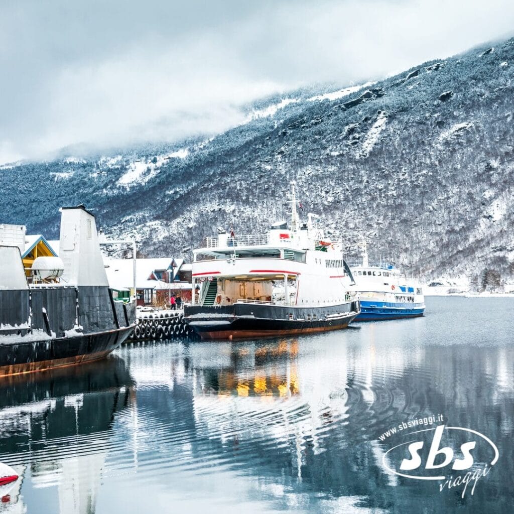 Traghetti attraccati in un porto innevato con montagne sullo sfondo e acque calme che riflettono la scena; logo "sbs viaggi" in primo piano, perfetto per un Tour con accompagnatore tra i Fiordi Meravigliosi.