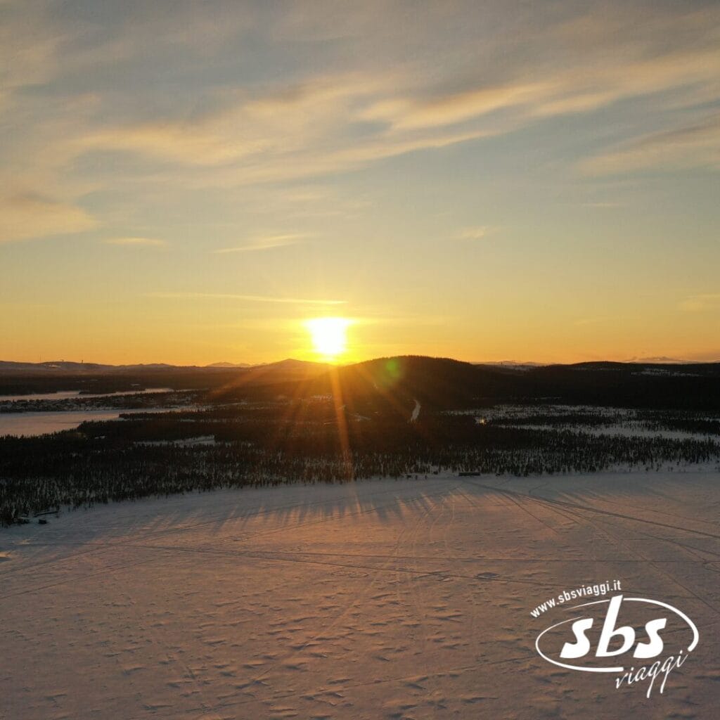 Il sole tramonta su un paesaggio innevato della Lapponia Svedese, con foreste e colline in lontananza; il logo di SBS Viaggi è visibile nell'angolo in basso a destra.