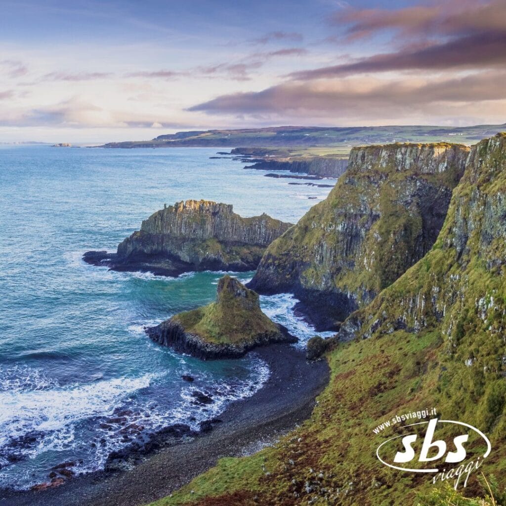 Tour Esclusivo Irlanda: Scogliere rocciose con erba verde si affacciano sul mare azzurro sotto un cielo parzialmente nuvoloso; le onde si infrangono lungo la costa frastagliata. Il logo