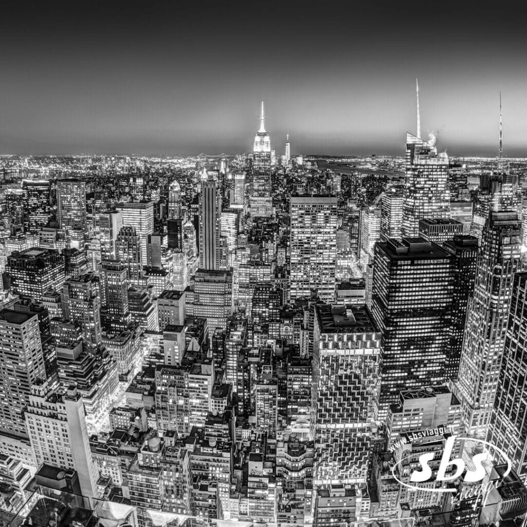 Vista aerea in bianco e nero di un denso skyline cittadino con alti grattacieli, luci brillanti e l'Empire State Building sullo sfondo centrale: perfetta per chi ha in programma di visitare New York con un tour esclusivo.