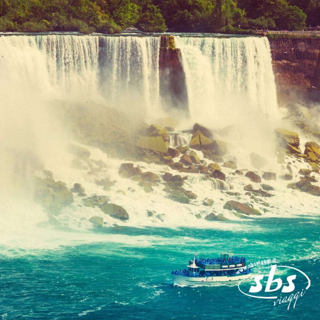 Un battello turistico pieno di persone si avvicina alla base della grande cascata del Niagara, circondato da nebbia, alberi e terreno roccioso. Il logo