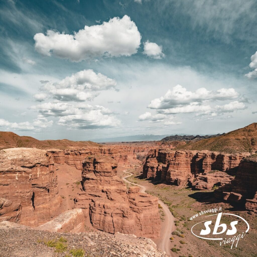 Una strada tortuosa attraversa un profondo canyon con formazioni rocciose rossastre sotto un cielo punteggiato di nuvole, catturando l'equilibrio tra tradizione e modernità; nell'angolo in basso a destra appare il logo di SBS Viaggi.