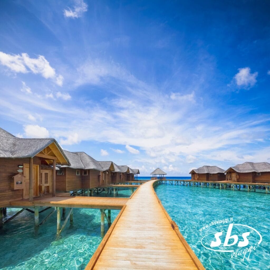 Passerella in legno sulle acque cristalline che conduce ai bungalow sull'acqua alle Maldive, sotto un cielo luminoso con nuvole sparse; logo 