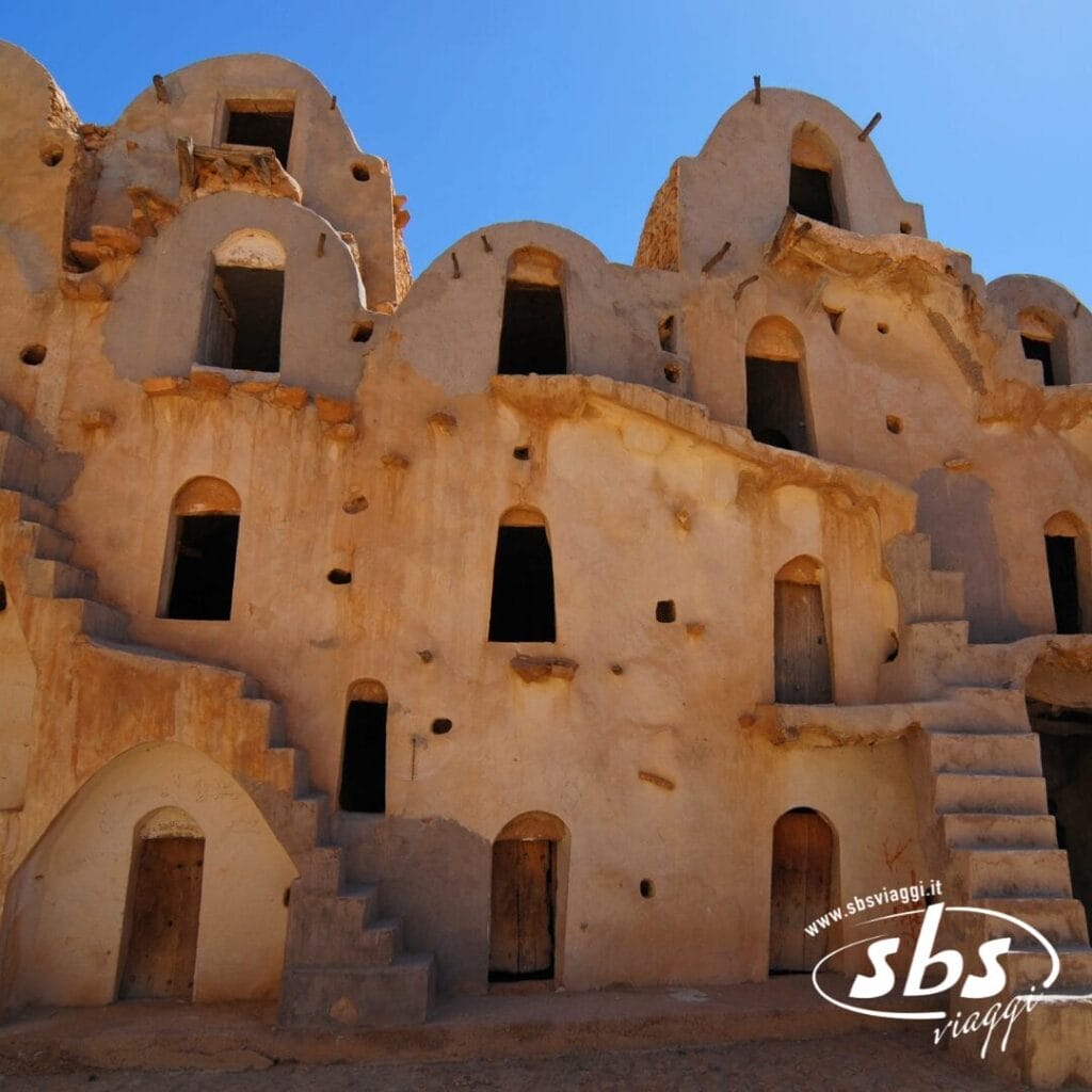Un antico edificio in terra battuta con porte e finestre ad arco, scale esterne, sotto un cielo azzurro e terso: uno spettacolo autentico nei pressi di un'oasi in Tunisia, perfetto per gli avventurieri in fuoristrada che esplorano il fascino unico della regione.