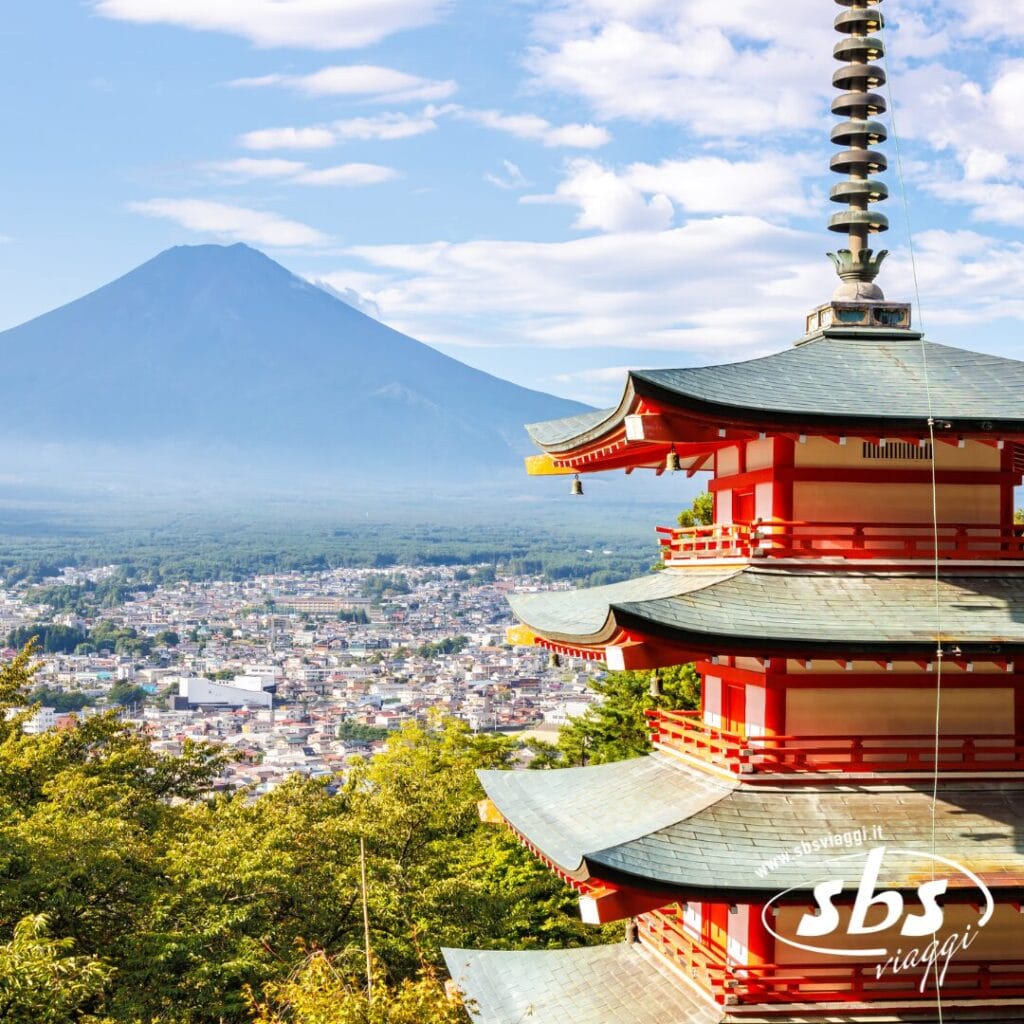 Pagoda rossa con una città e il Monte Fuji sullo sfondo, sotto un cielo parzialmente nuvoloso, che cattura lo spirito del Giappone. Il logo di SBS Viaggi è visibile nell'angolo in basso a destra.