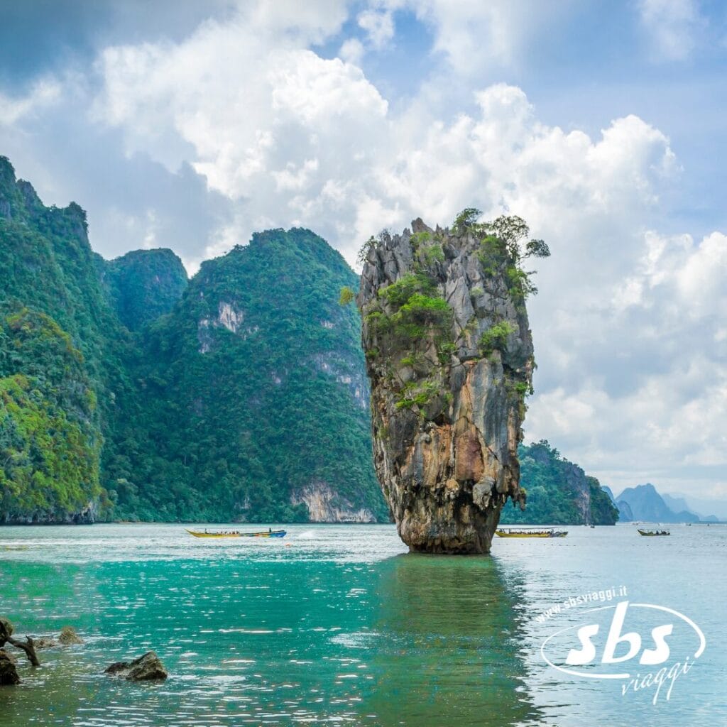 Un'alta isola rocciosa ricoperta di verde si erge dalle acque turchesi, con le montagne sullo sfondo, catturando la magia della Thailandia Magica e la serena bellezza vicino a Phuket.