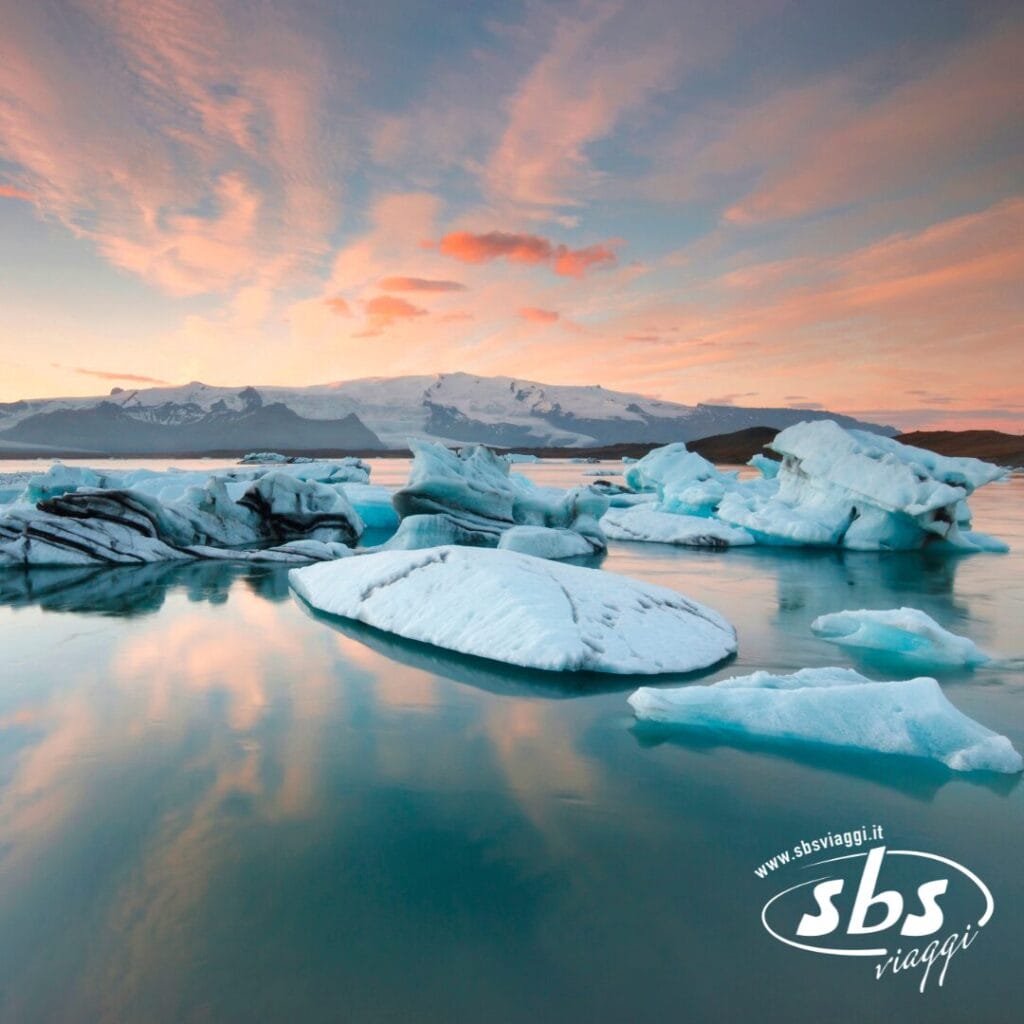 Iceberg che galleggiano in acque calme al tramonto in Islanda, con montagne innevate e un cielo colorato sullo sfondo.