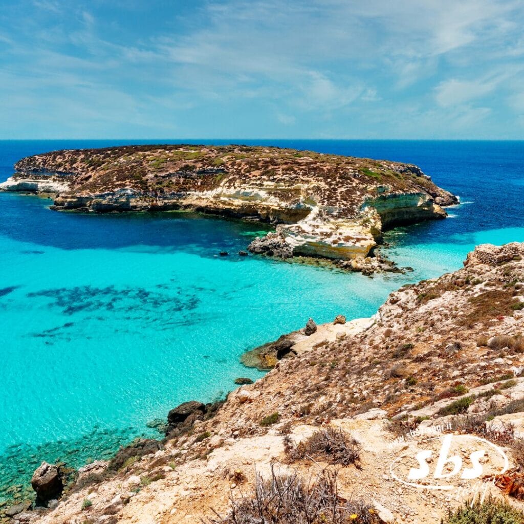 Un'isola rocciosa circondata da acque turchesi e cristalline sotto un cielo azzurro brillante, con vegetazione secca in primo piano: una vista idilliaca vicino a Case Vacanza Lampedusa.