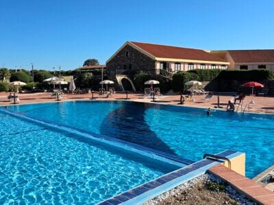 Ampia piscina all'aperto con acqua cristallina, sedie a sdraio e ombrelloni vicino a un edificio con il tetto rosso: perfetta per rilassarsi in questa cornice di Bozza automatica.