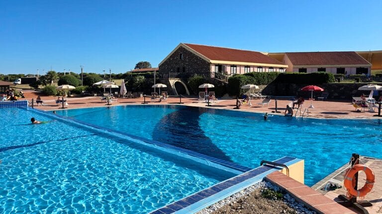 Ampia piscina all'aperto con acqua cristallina, sedie a sdraio e ombrelloni vicino a un edificio con il tetto rosso: perfetta per rilassarsi in questa cornice di Bozza automatica.