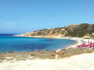 Una spiaggia sabbiosa con acqua turchese, colline rocciose e file di sedie a sdraio rosse sotto gli ombrelloni creano il luogo perfetto per una fuga a Bozza automatica.