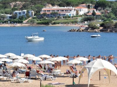 Persone che si tengono per mano in cerchio su una spiaggia sabbiosa, con ombrelloni, barche sull'acqua e case sullo sfondo, creano una scena vivace che ricorda una bozza automatica.