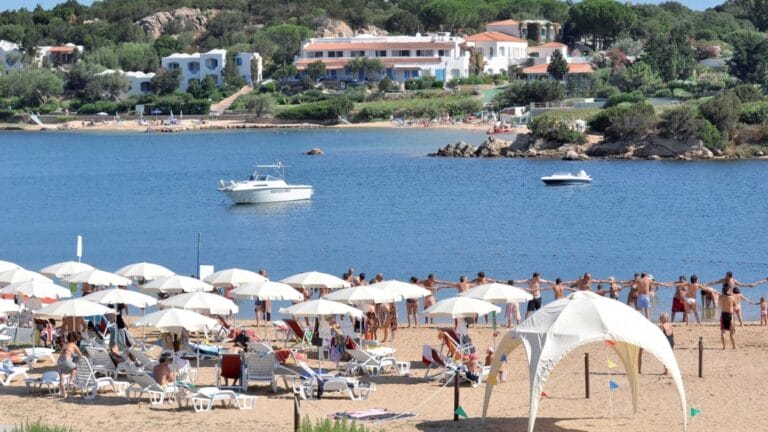 Persone che si tengono per mano in cerchio su una spiaggia sabbiosa, con ombrelloni, barche sull'acqua e case sullo sfondo, creano una scena vivace che ricorda una bozza automatica.