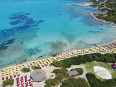 Vista aerea della spiaggia di Stintino con acqua turchese, ombrelloni gialli e bagnanti lungo la riva vicino al Club Esse Roccaruja.