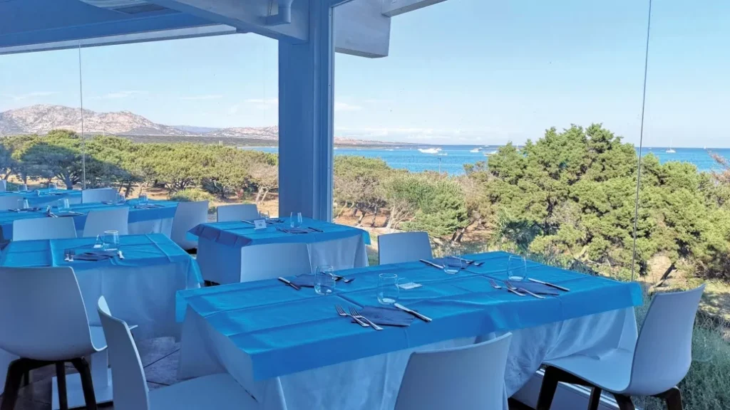 Ristorante con tovaglie blu, sedie bianche e ampie vetrate con vista sul mare e sugli alberi in una giornata di sole al Club Esse Roccaruja di Stintino.