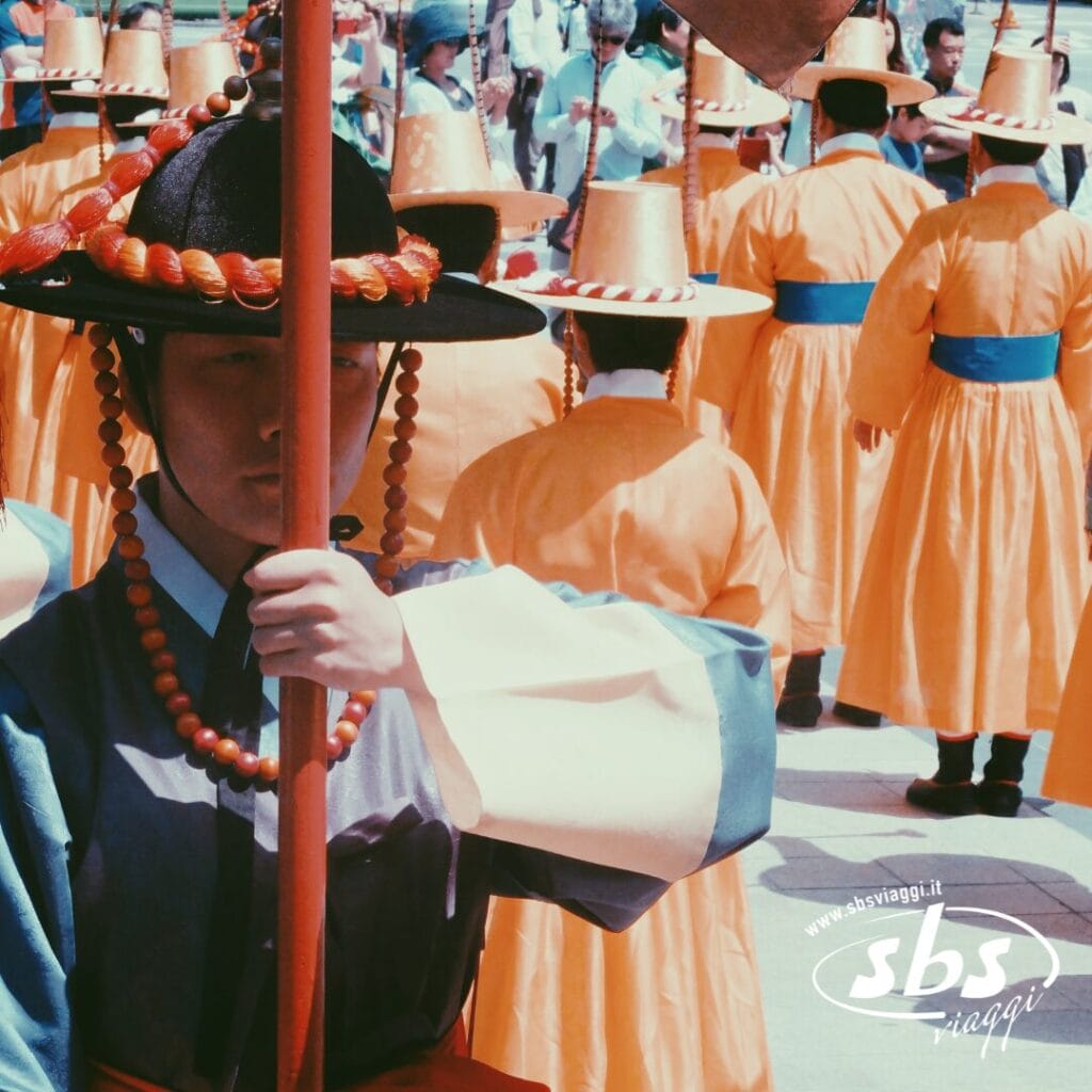 Un gruppo di persone in abiti tradizionali coreani partecipa a una sfilata durante il Tour Corea del Sud; una persona in testa tiene un palo e indossa una collana di perline e un cappello a tesa larga.