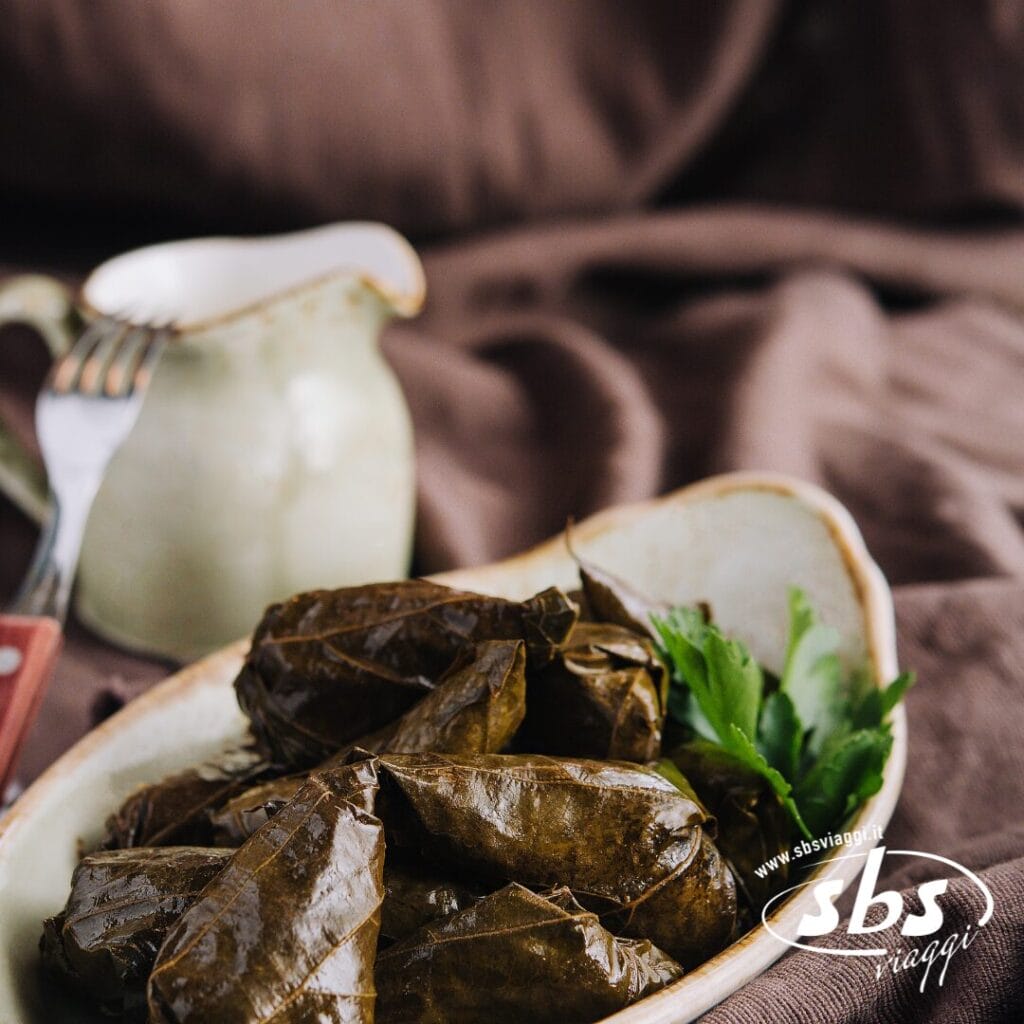 Un piatto di ceramica con foglie di vite ripiene, un piatto popolare in Azerbaigian e Georgia, guarnito con prezzemolo, con una piccola brocca e una forchetta sullo sfondo.