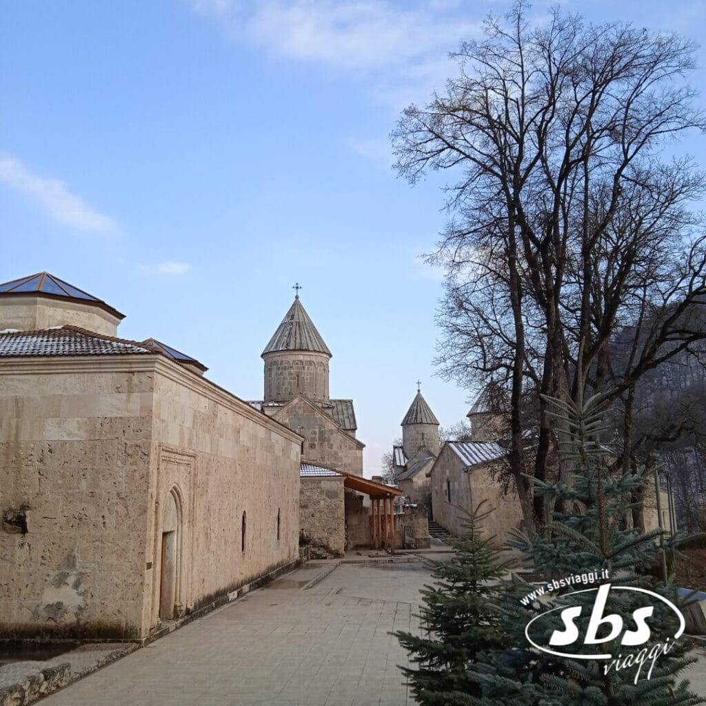 Edifici in pietra e campanili con tetto conico, tipici dell'Armenia e della Georgia, costeggiano un sentiero lastricato, con alberi spogli e arbusti sempreverdi visibili sotto un cielo parzialmente nuvoloso.
