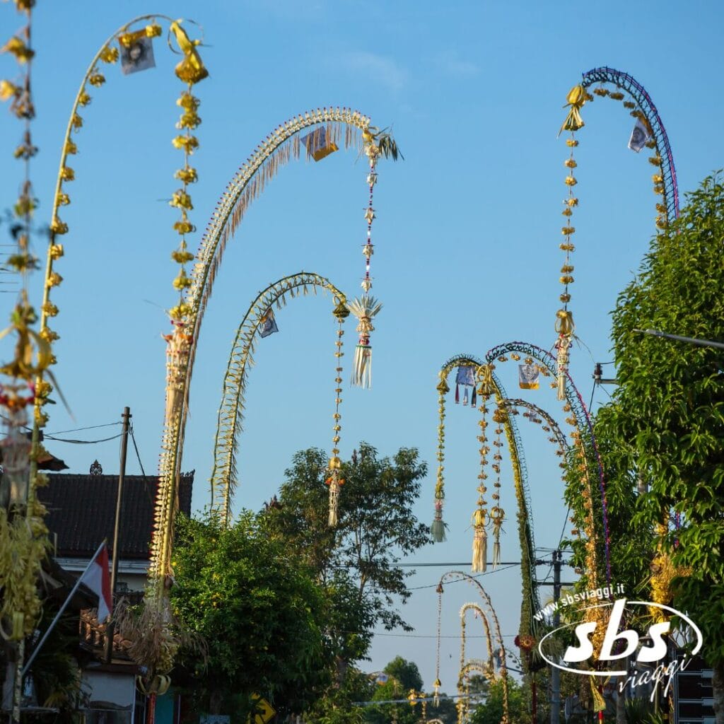 Alti pali di bambù decorati costeggiano una strada sotto un cielo azzurro e terso, da cui pendono ornamenti tradizionali e fogliame: un momento Instagram perfetto per chi ama esplorare la vivace cultura di Bali.