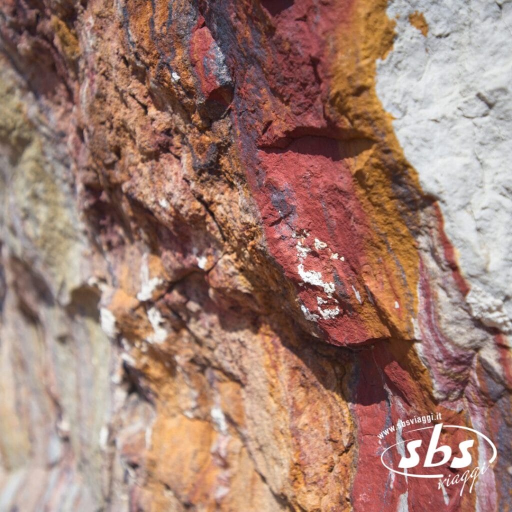 Primo piano della superficie di una roccia caratterizzata da strati di minerali rossi, gialli e bianchi nella Valle degli Dei in Nepal, con il logo