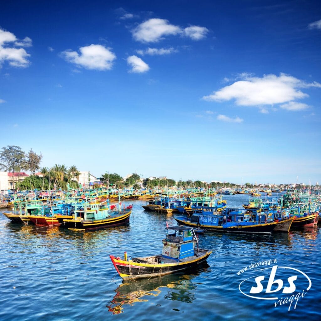 Colorate barche da pesca sono ormeggiate in un porto in una giornata di sole, con cielo azzurro e nuvole sparse. Il logo di SBS Viaggi è visibile nell'angolo, evocando lo spirito vibrante del tour Vietnam Classico.