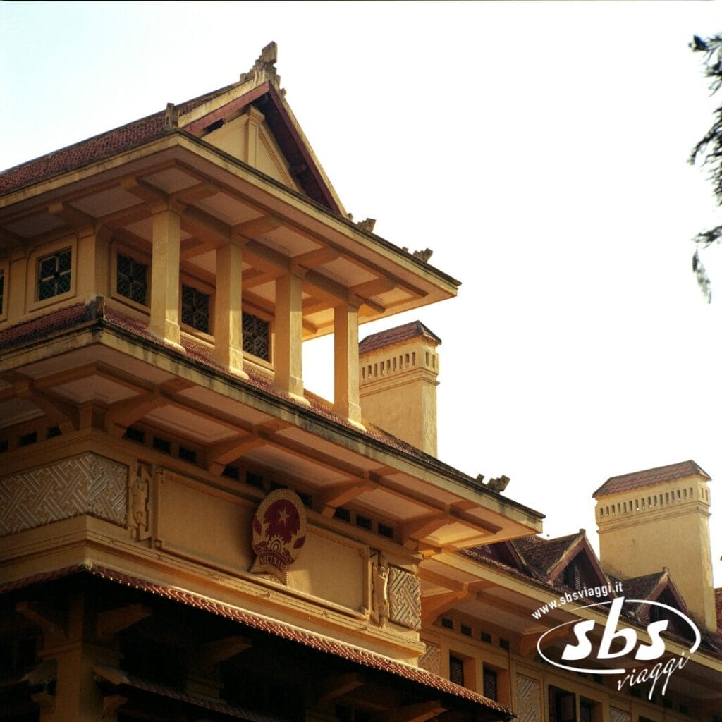 Un edificio giallo con dettagli architettonici tradizionali, con l'emblema nazionale vietnamita sopra l'ingresso, offre un'esperienza unica in Vietnam sotto un cielo limpido.