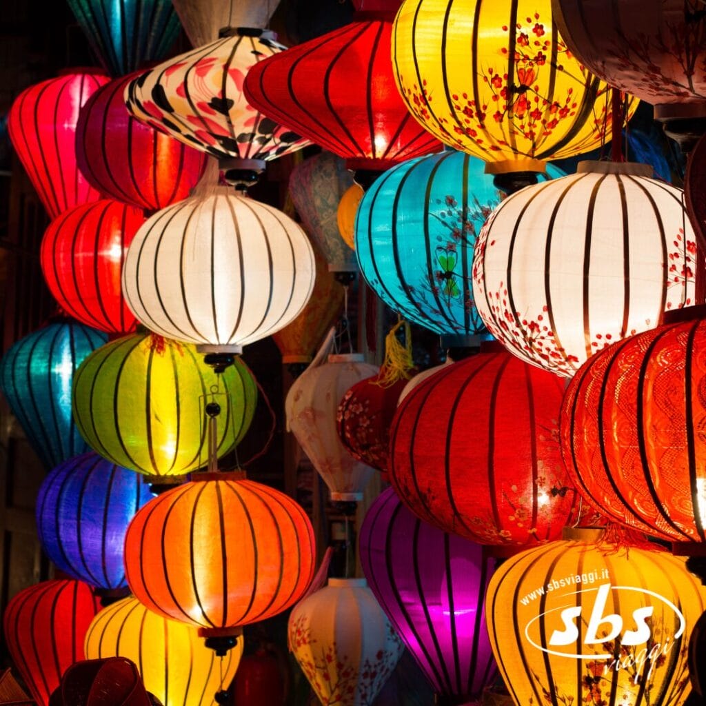 Lanterne colorate di varie forme e fantasie sono appese vicine, illuminate e creano un'atmosfera vivace: una scena iconica che si ritrova spesso durante il Gran Tour del Vietnam. L'immagine presenta la filigrana