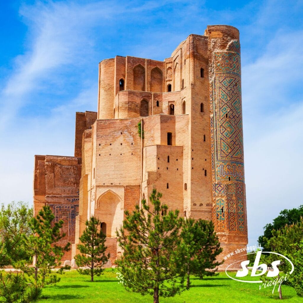 Un grande e antico edificio in mattoni con intricate piastrelle si erge circondato da prati verdi e piccoli pini sotto un cielo azzurro: uno scenario senza tempo dell'Uzbekistan, perfetto per qualsiasi itinerario Grantour.