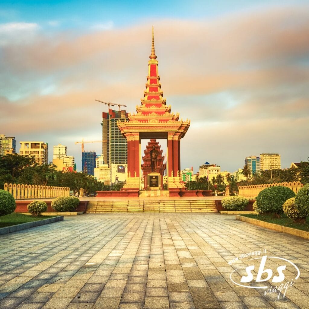 Un monumento con il tetto rosso e oro si erge in una piazza, circondato dal verde, con edifici moderni e gru sullo sfondo: un luogo suggestivo da visitare durante la tua avventura con Grantour o Tour Cambogia.