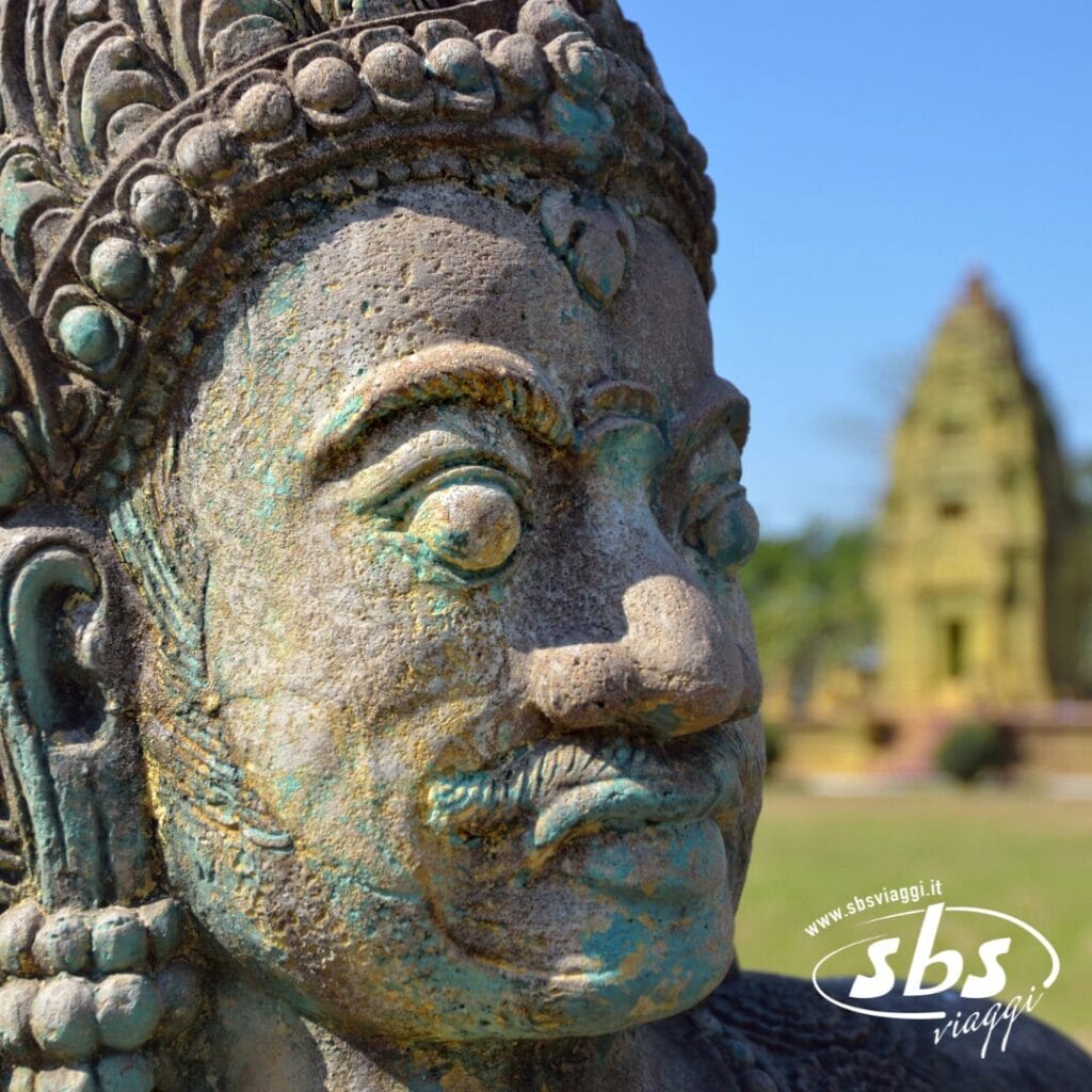 Primo piano di una statua in pietra erosa di una divinità con un elaborato copricapo, con un antico tempio sfocato sullo sfondo: tutto ciò cattura lo spirito di una vera esperienza Tour Cambogia.