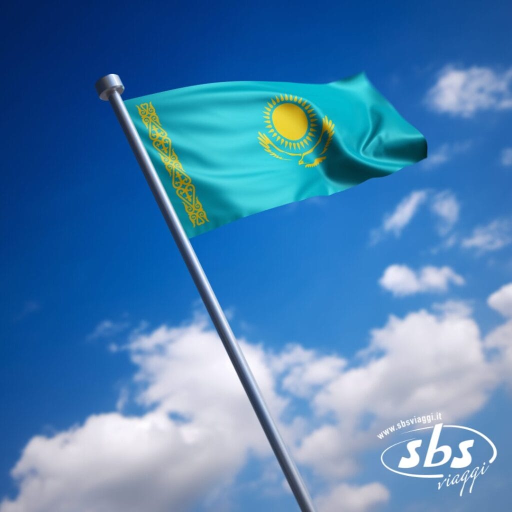 La bandiera del Kazakistan, raffigurante un sole, un'aquila e un motivo ornamentale su sfondo blu, sventola su un pennone contro un cielo parzialmente nuvoloso: uno spettacolo suggestivo per chiunque abbia in programma di visitare il Kazakistan.