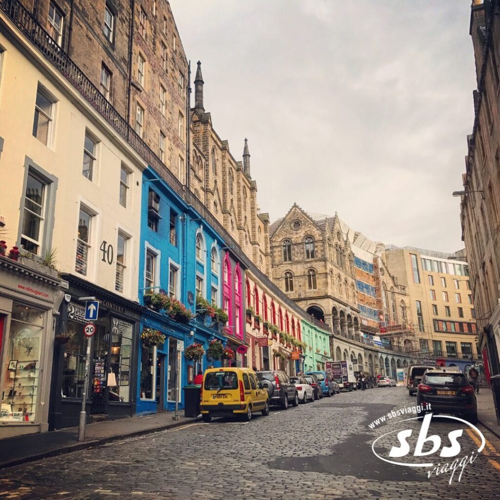Una strada acciottolata fiancheggiata da edifici colorati, auto parcheggiate e pedoni evoca il fascino di un Tour Scozia, con l'architettura storica delle Highlands sullo sfondo sotto un cielo nuvoloso.