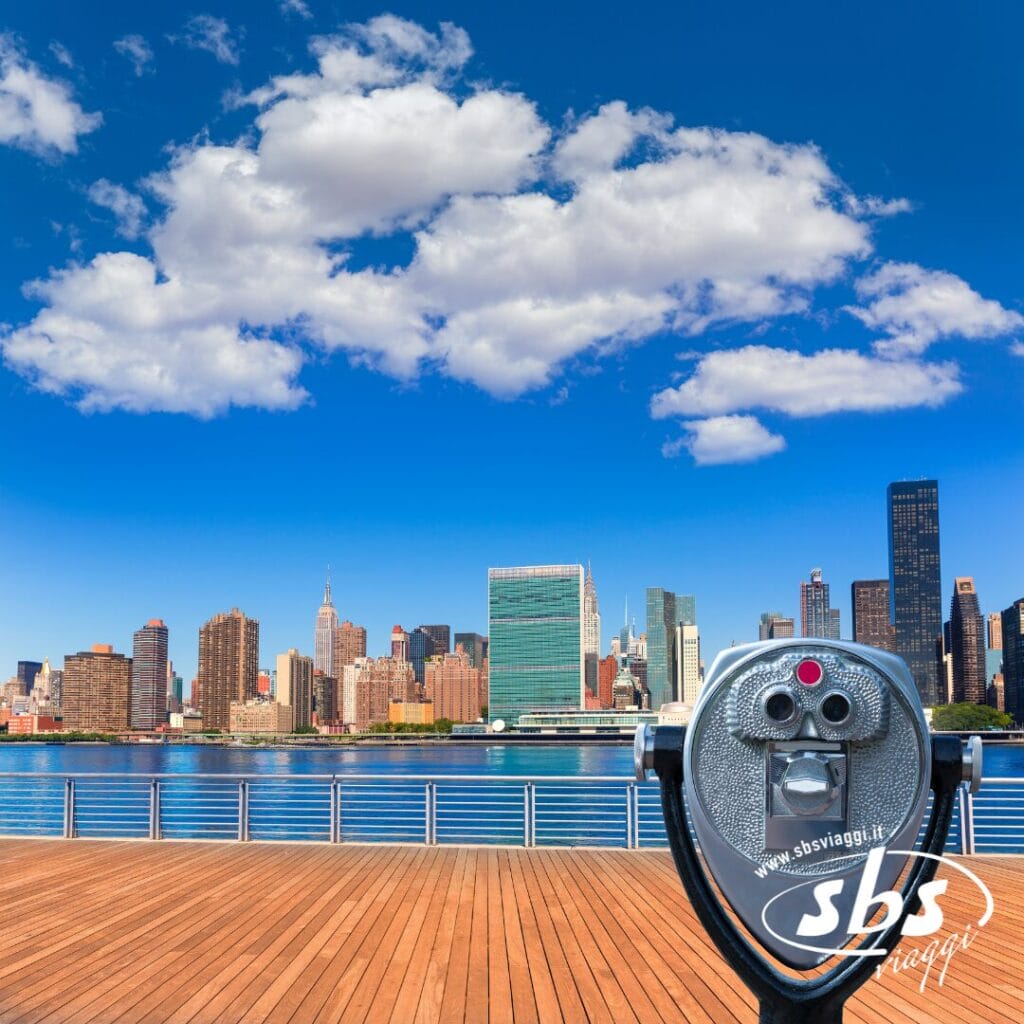 Un visore a gettoni su una piattaforma di legno domina lo skyline di New York City sotto un cielo azzurro con nuvole sparse, catturando la magia di Settembre a New York: perfetto per un accompagnatore dall'Italia in cerca di viste mozzafiato.