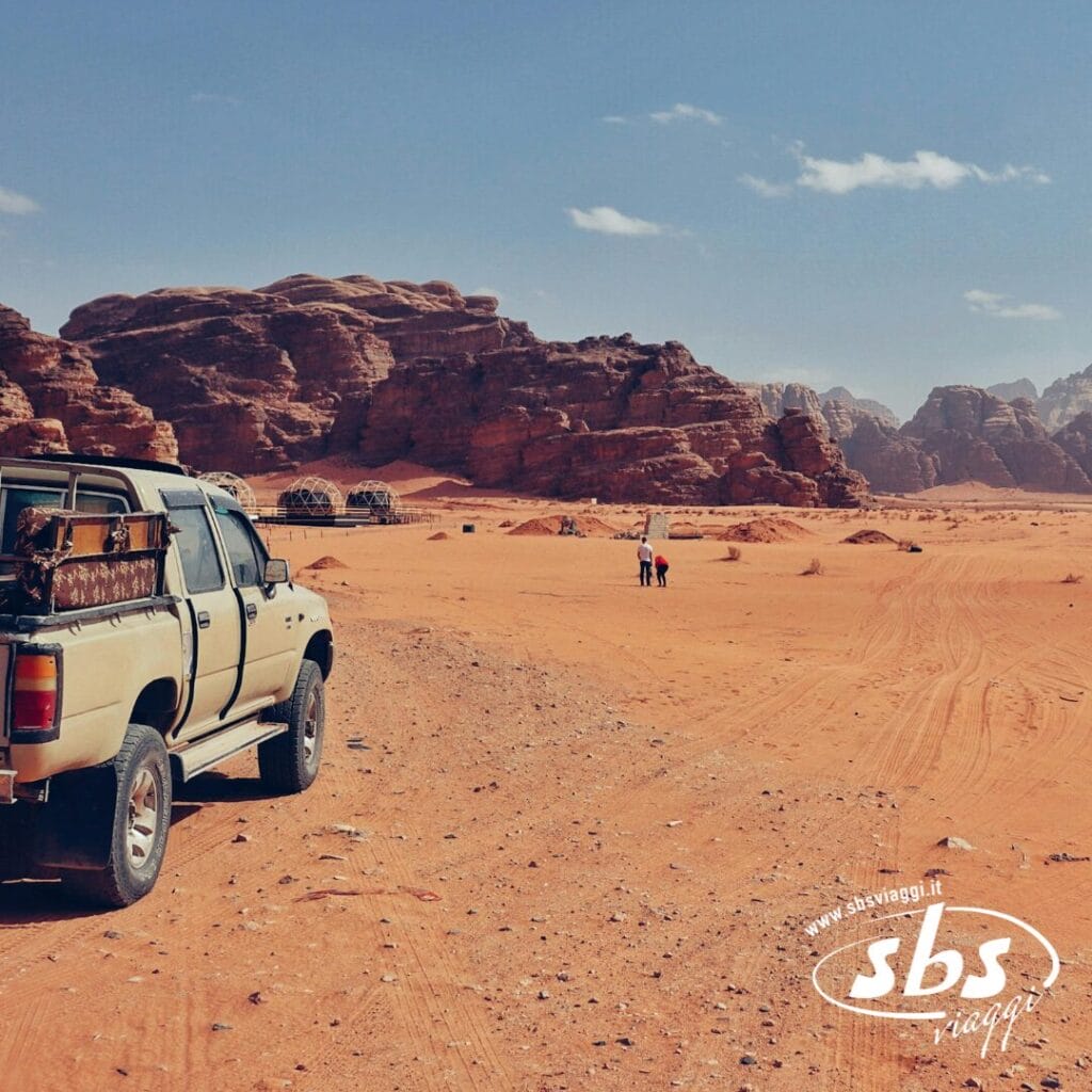 Un pick-up beige in un paesaggio desertico sabbioso con grandi formazioni rocciose, dove lo spirito della Tradizione incontra la Modernità del viaggio, e due persone camminano in lontananza.