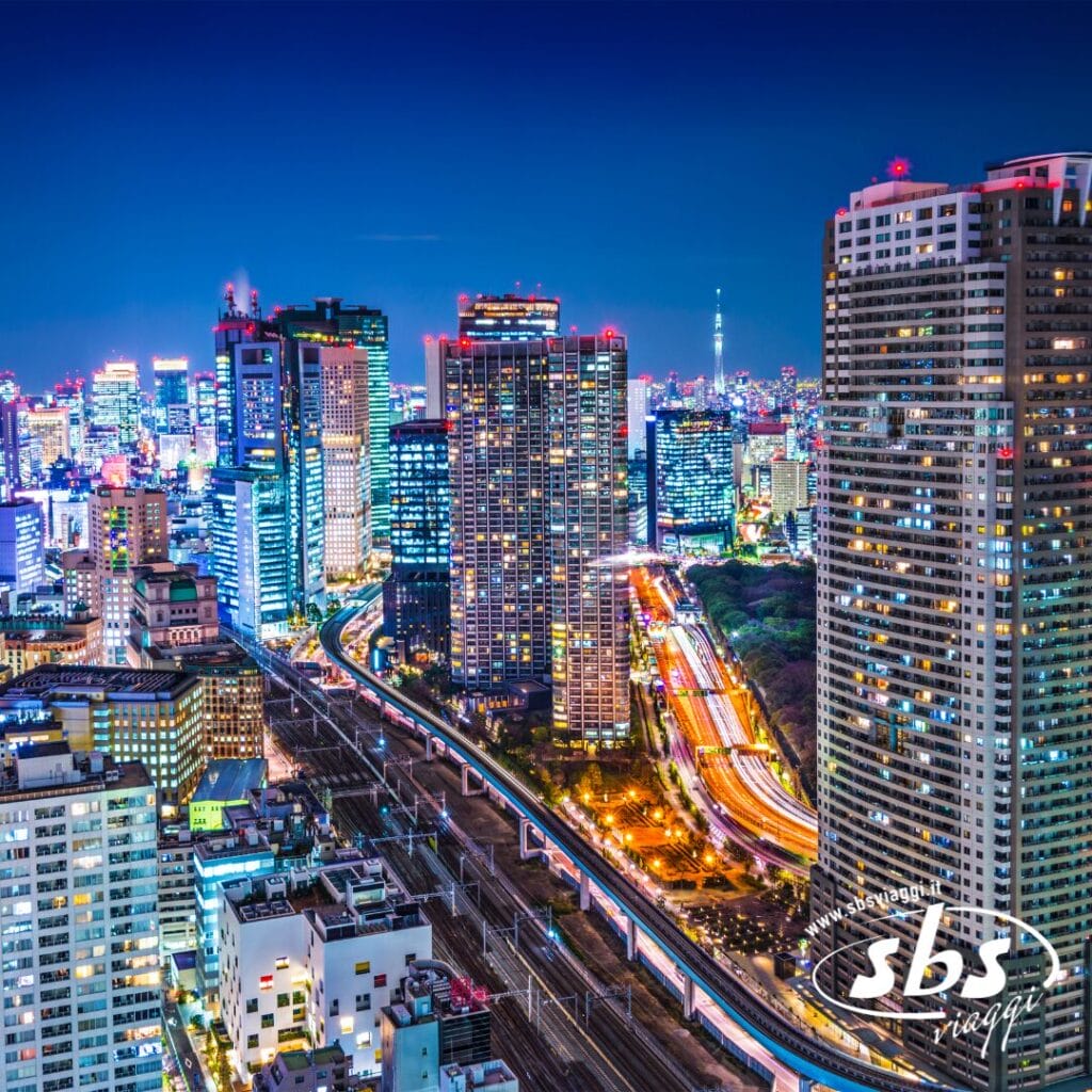 Paesaggio urbano notturno con alti edifici, strade illuminate e una carreggiata visibile con scie luminose del traffico in Giappone; logo