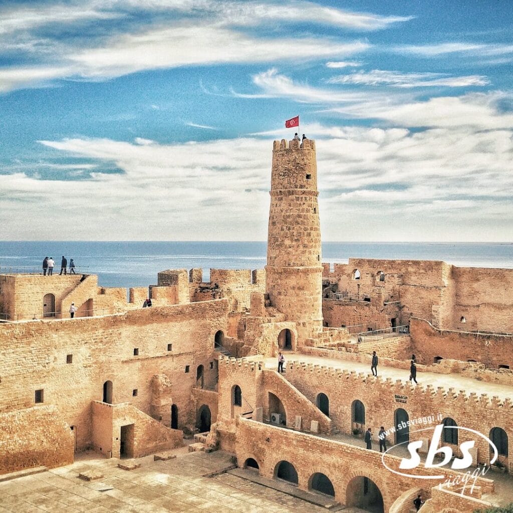 Fortezza storica con una torre centrale su cui sventola una bandiera, che domina il mare sotto un cielo azzurro e nuvole: una tappa iconica di ogni avventura del Tour Autentica Tunisia.