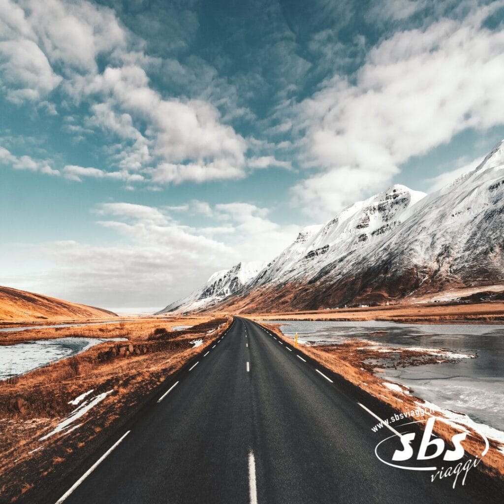 Strada aperta che conduce verso montagne innevate sotto un cielo parzialmente nuvoloso, con acqua su entrambi i lati: un invito alla scoperta tra meravigliosi altopiani.