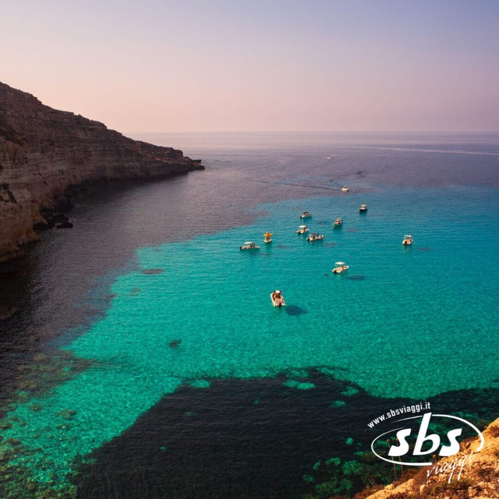Piccole imbarcazioni galleggiano sulle limpide acque turchesi vicino alle scogliere rocciose, sotto un cielo velato, con il logo "sbs viaggi" e Case Vacanza Lampedusa ben visibili.