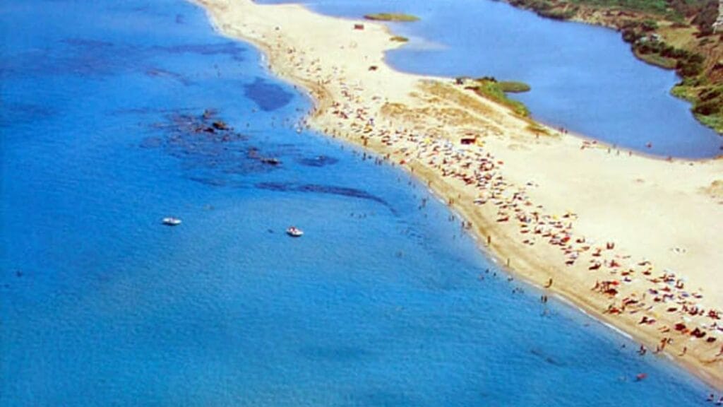 Una spiaggia sabbiosa con acqua turchese, folle di persone che prendono il sole e una piccola laguna nelle vicinanze creano uno scenario vivace, perfetto per una Bozza automatica.