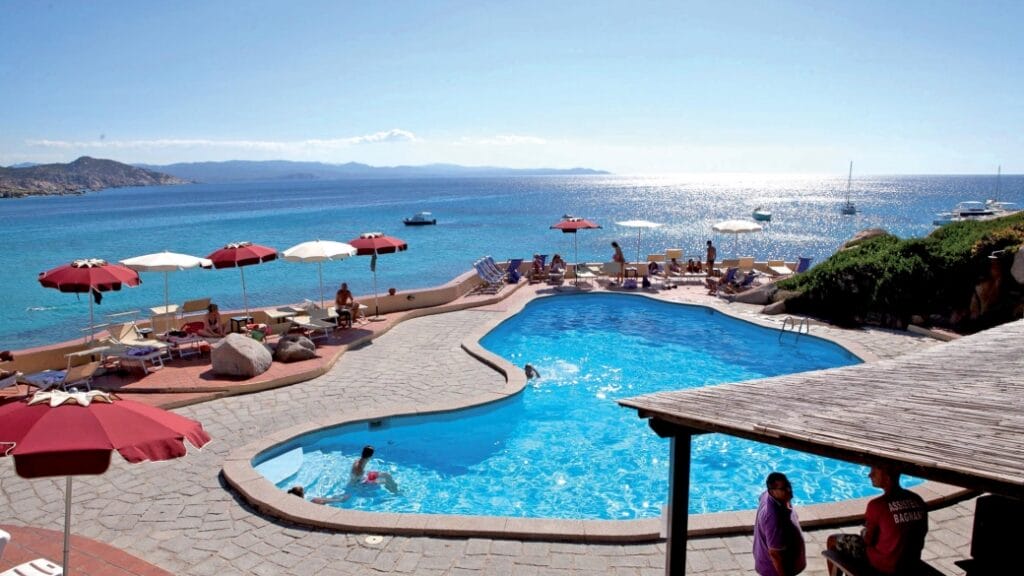 Una piscina di un resort sul mare con ombrelloni rossi, sedie a sdraio e persone che si rilassano in riva all'oceano blu offre la fuga perfetta: un ambiente invitante in cui rilassarsi con stile.