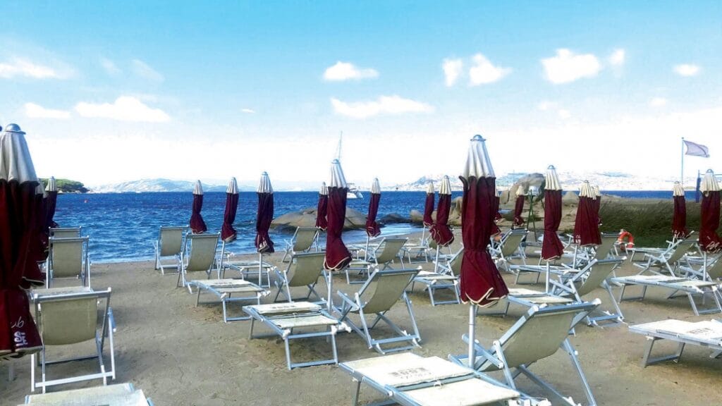 Sedie a sdraio vuote e ombrelloni chiusi costeggiano la riva sabbiosa sotto un cielo e un mare azzurri e limpidi, catturando un sereno momento di quiete, come una perfetta bozza automatica in attesa del tocco finale.