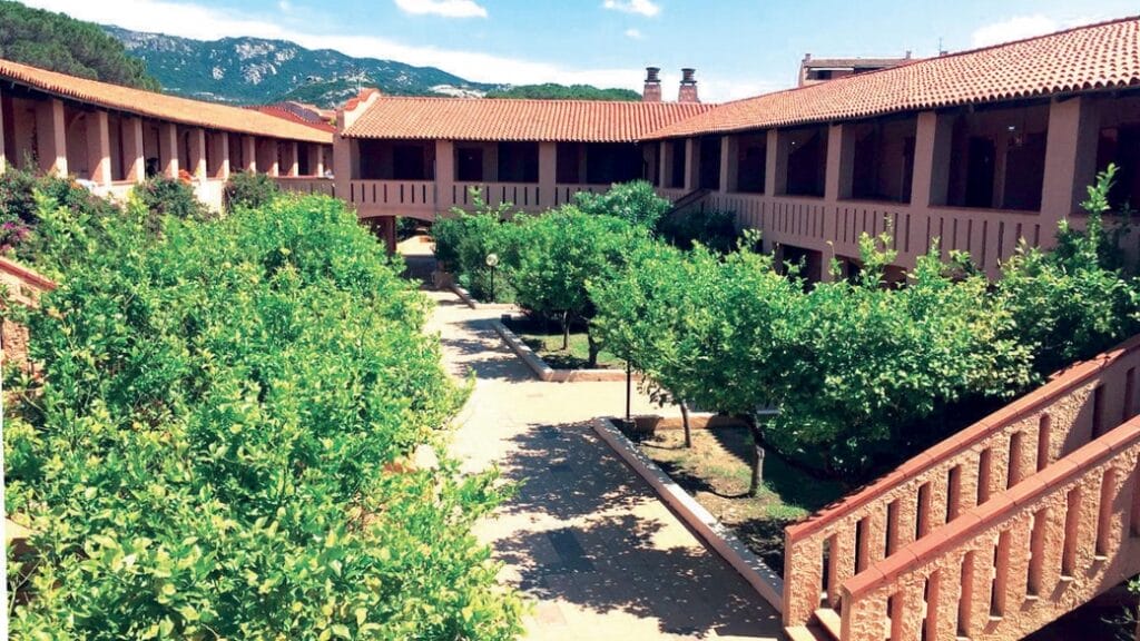 Cortile con rigogliosi alberi verdi e Bozza automatica, circondato da un edificio a due piani con tetto in tegole rosse e montagne sullo sfondo.