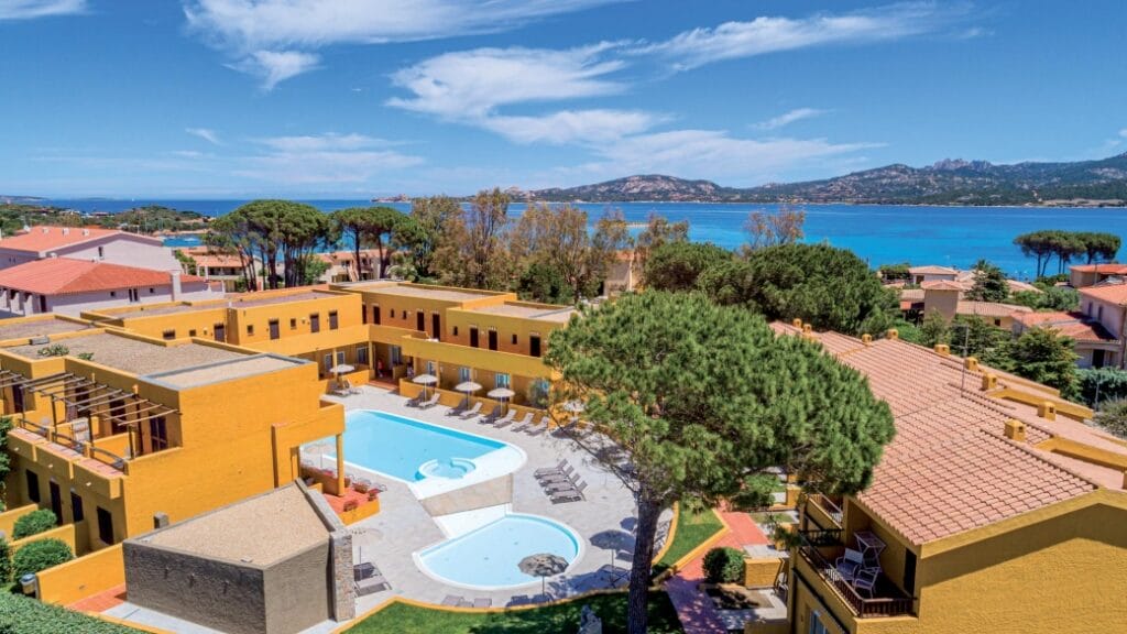 Vista aerea di un resort giallo con piscina vicino al mare, circondato da alberi e montagne sullo sfondo: un'invitante fuga alla Bozza automatica.