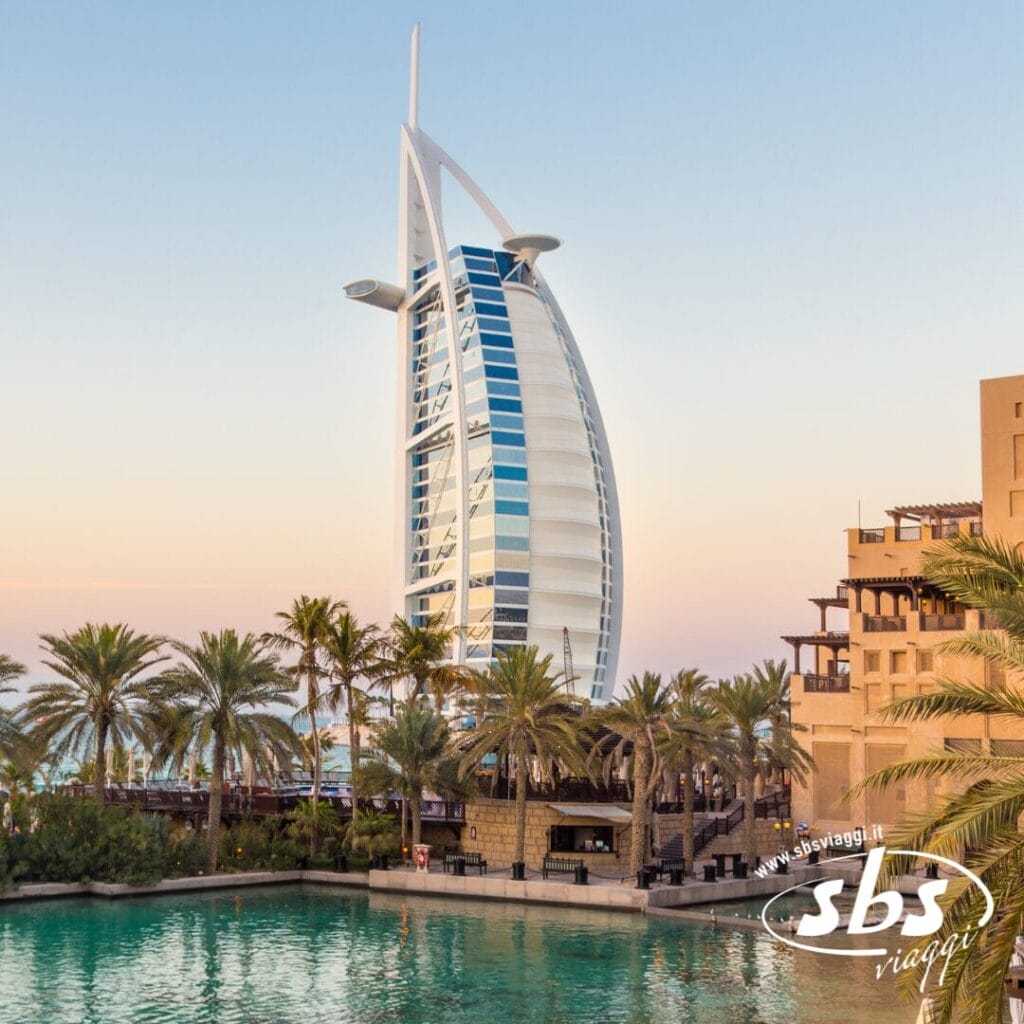 Il moderno hotel Burj Al Arab a forma di vela al tramonto, con palme e una piscina turchese in primo piano: una vista iconica che ricorda il lusso del Demo Hotel e l'eleganza del Demo Villaggi.
