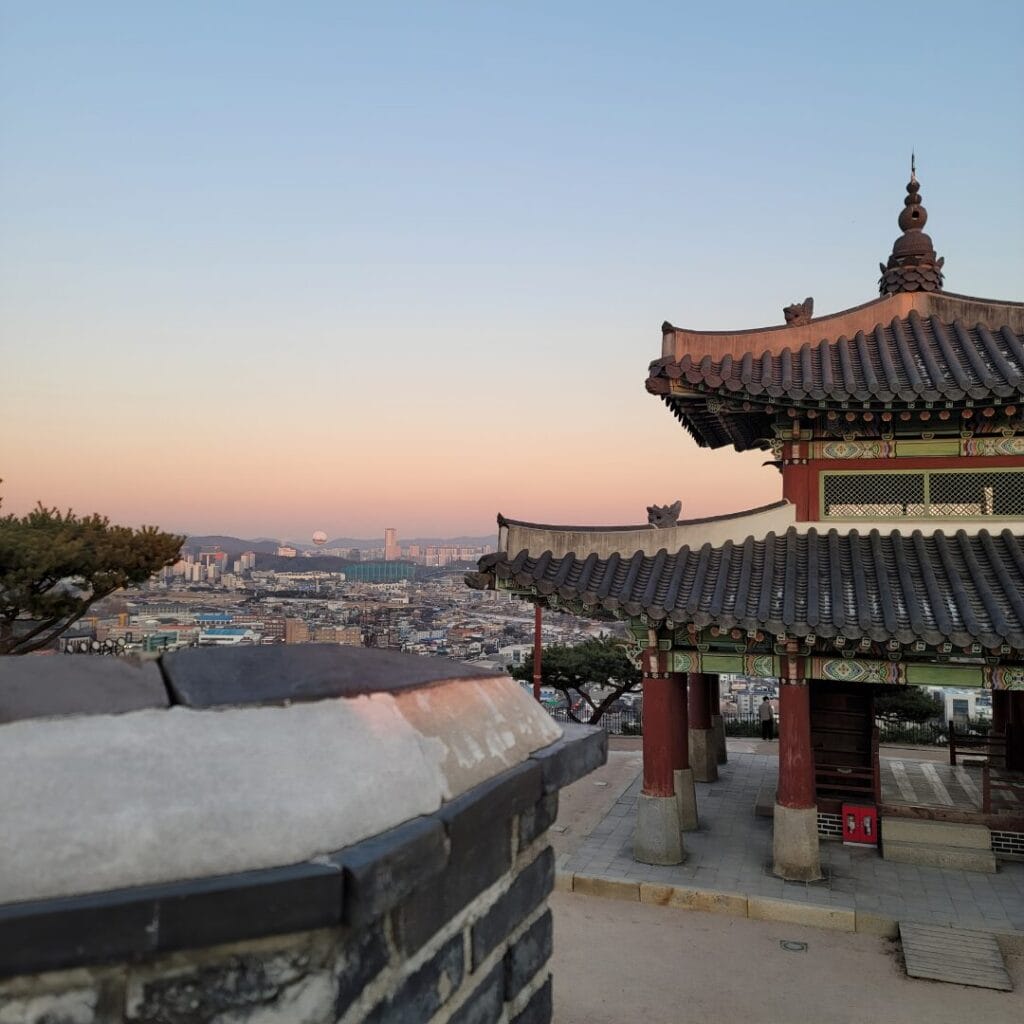 Un tradizionale padiglione coreano domina il paesaggio urbano al tramonto, con montagne ed edifici sullo sfondo: uno spettacolo indimenticabile durante il vostro Tour Corea del Sud sotto il cielo limpido.